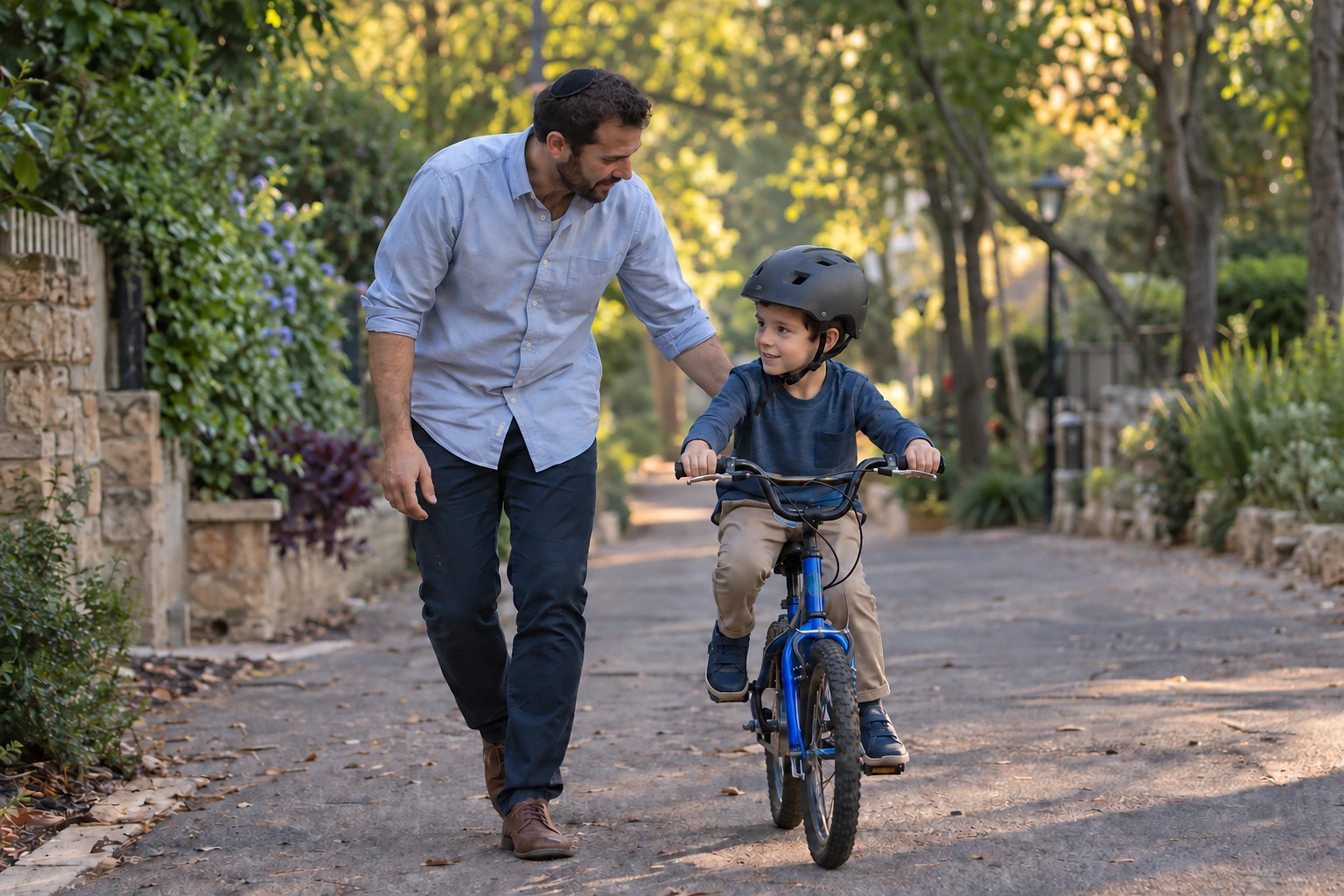 A father wearing a black kippah helping his helmeted son learn to ride a bicycle